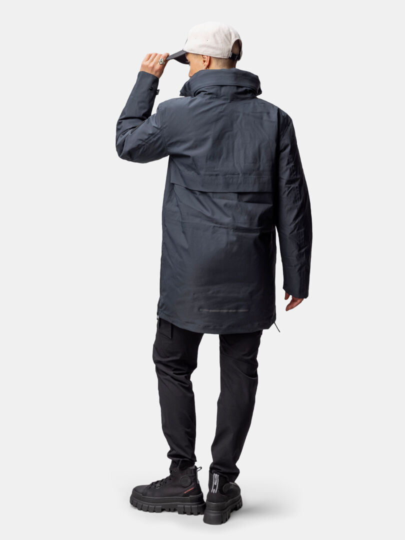 Henkilö, jolla on Halti Synkas 3in1 DrymaxX Parka, mustat housut ja kengät, säätää valomyssyä ja katsoo poispäin.