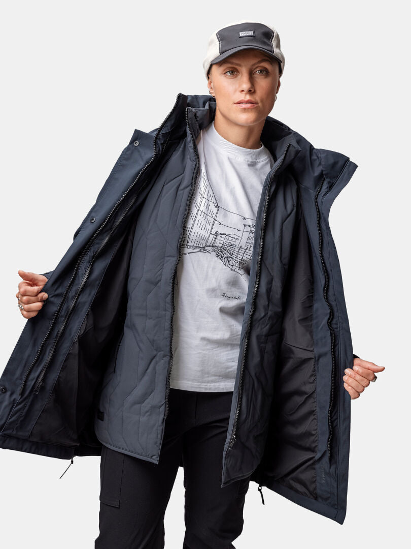 Henkilö pukeutuu graafiseen t-paitaan ja Halti Synkas 3in1 DrymaxX Parka Naisten -parka-asuun pitäen sitä auki, jotta sen sisus näkyy.