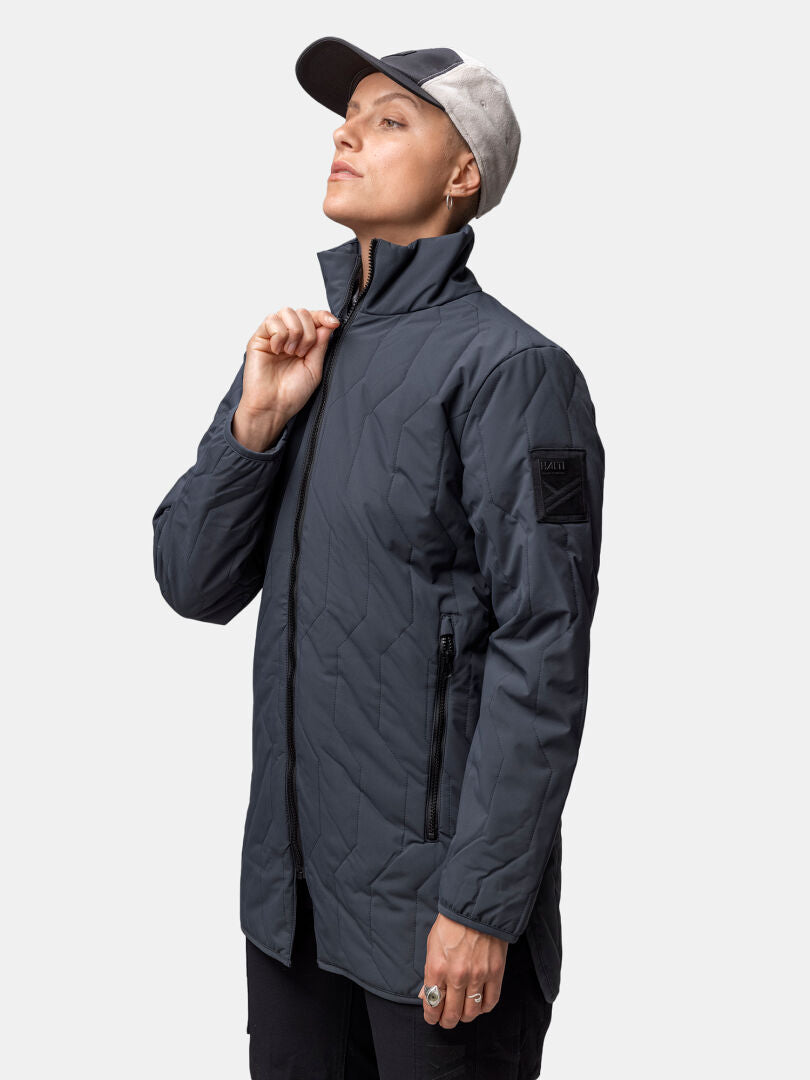 Henkilö, jolla on Halti Synkas 3in1 DrymaxX Parka ja kaksivärinen lippis, seisoo pitelemässä takin vetoketjua tavallista taustaa vasten.
