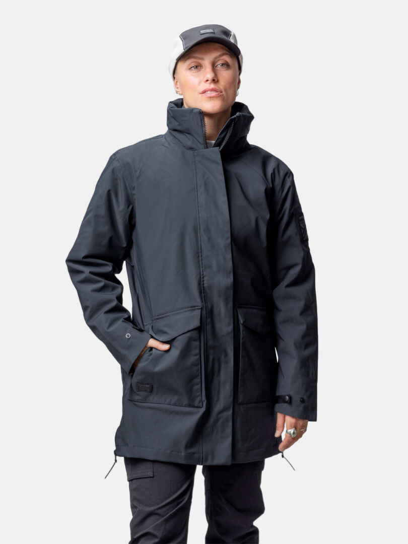 Henkilö, jolla on Halti Synkas 3in1 DrymaxX Parka, vaalea lippis ja mustat housut valkoista taustaa vasten.