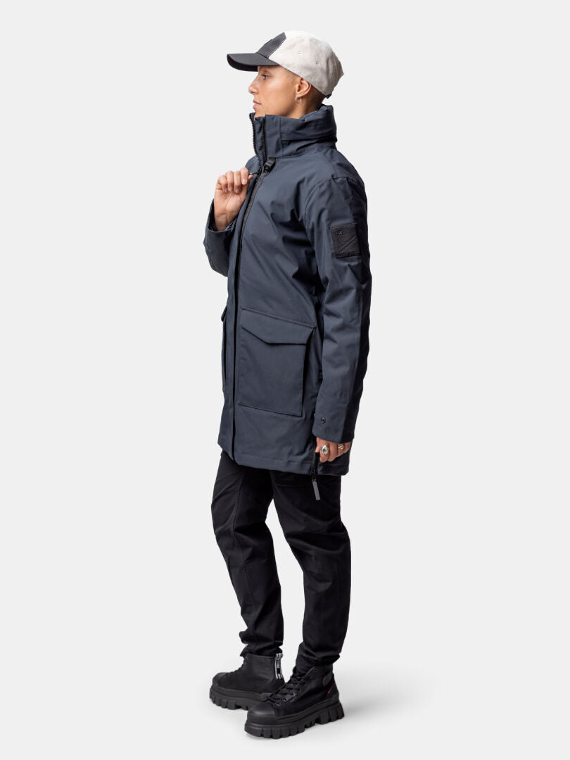 Henkilö, jolla on harmaa Halti Synkas 3in1 DrymaxX Parka, mustat housut, saappaat ja lippis, seisoo vasemmalle päin tavallista taustaa vasten.