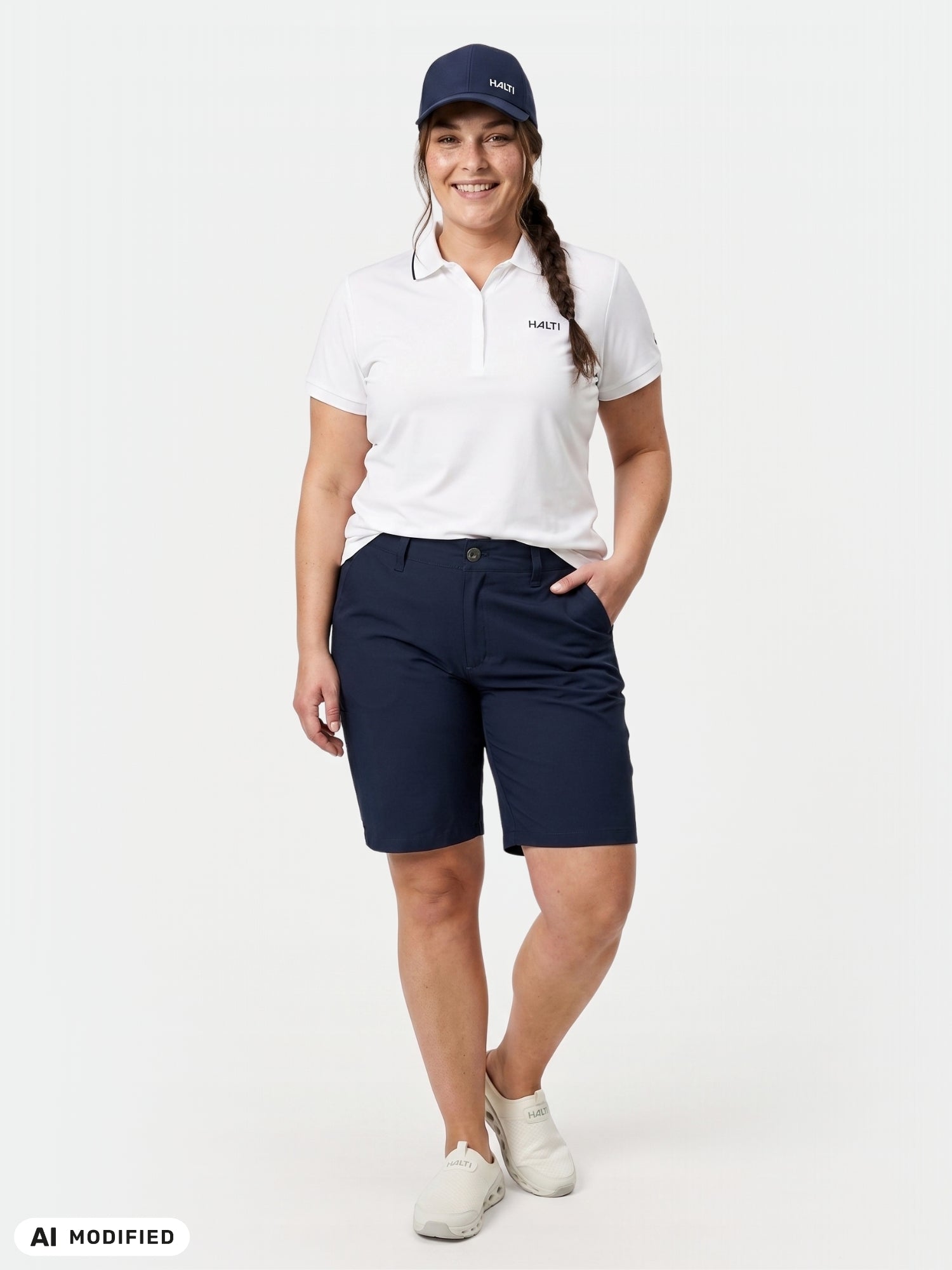 Nainen, jolla on yllään Halti Drive X-stretch Shortsit Naisten Plus -shortsit, valkoinen poolopaita ja lippalakki, seisoo käsi taskussa valkoista taustaa vasten.