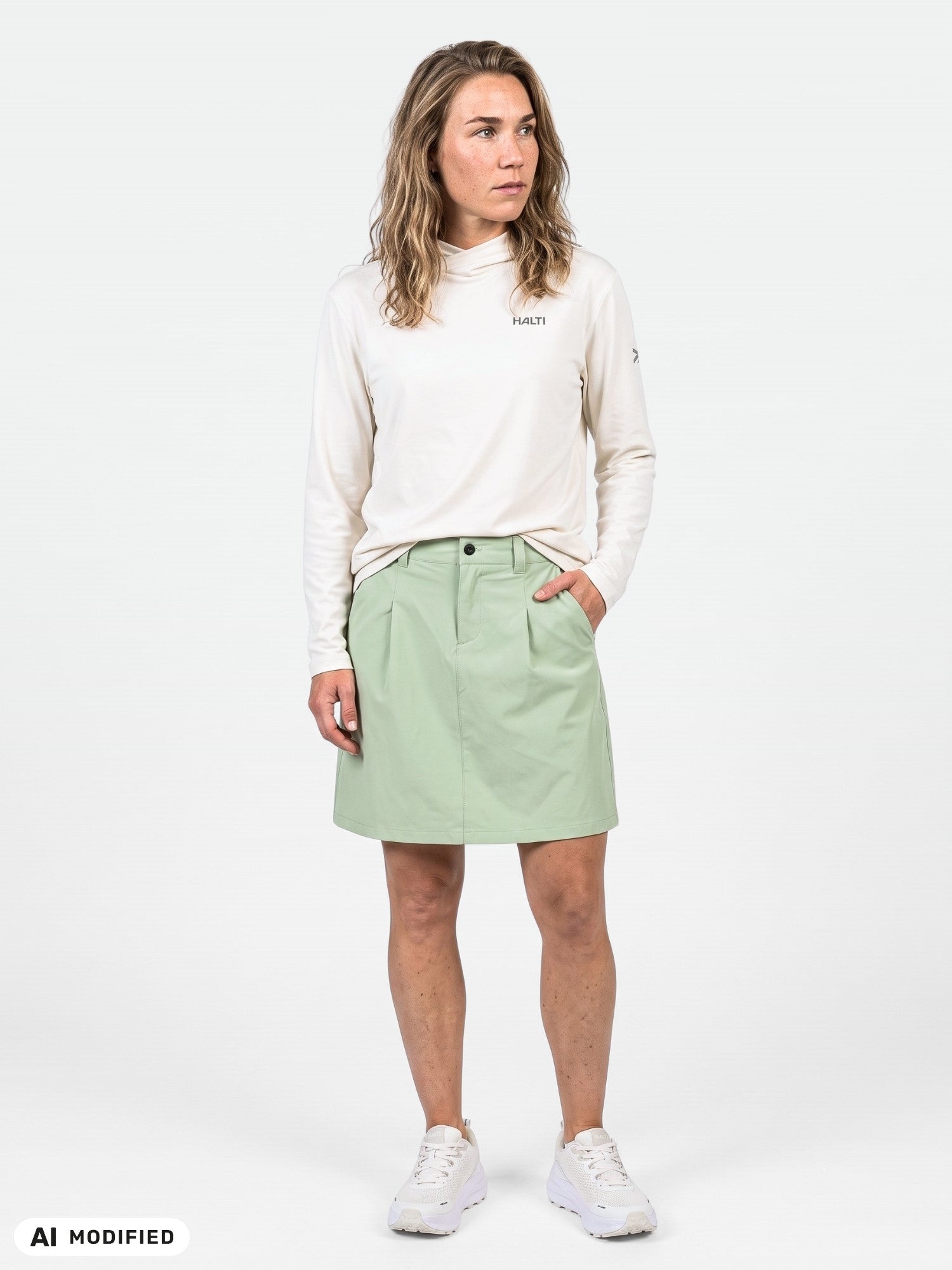 Henkilö, jolla on yllään valkoinen pitkähihainen paita, Halti Drive X-stretch Shortsihame Naisten -shortsit ja valkoiset lenkkarit, tasaisella taustalla.
