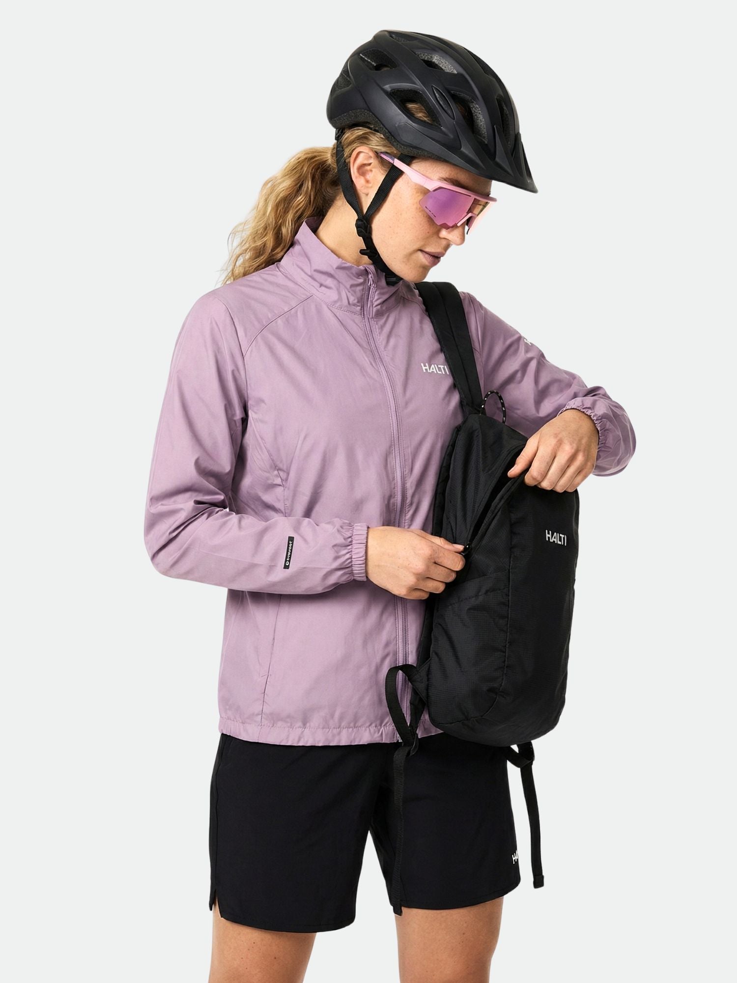 Kypärä ja aurinkolasit päässä oleva nainen pukeutuu Halti Saime Tuulitakki Naisten ja mustiin shortseihin ja avaa mustan repun.