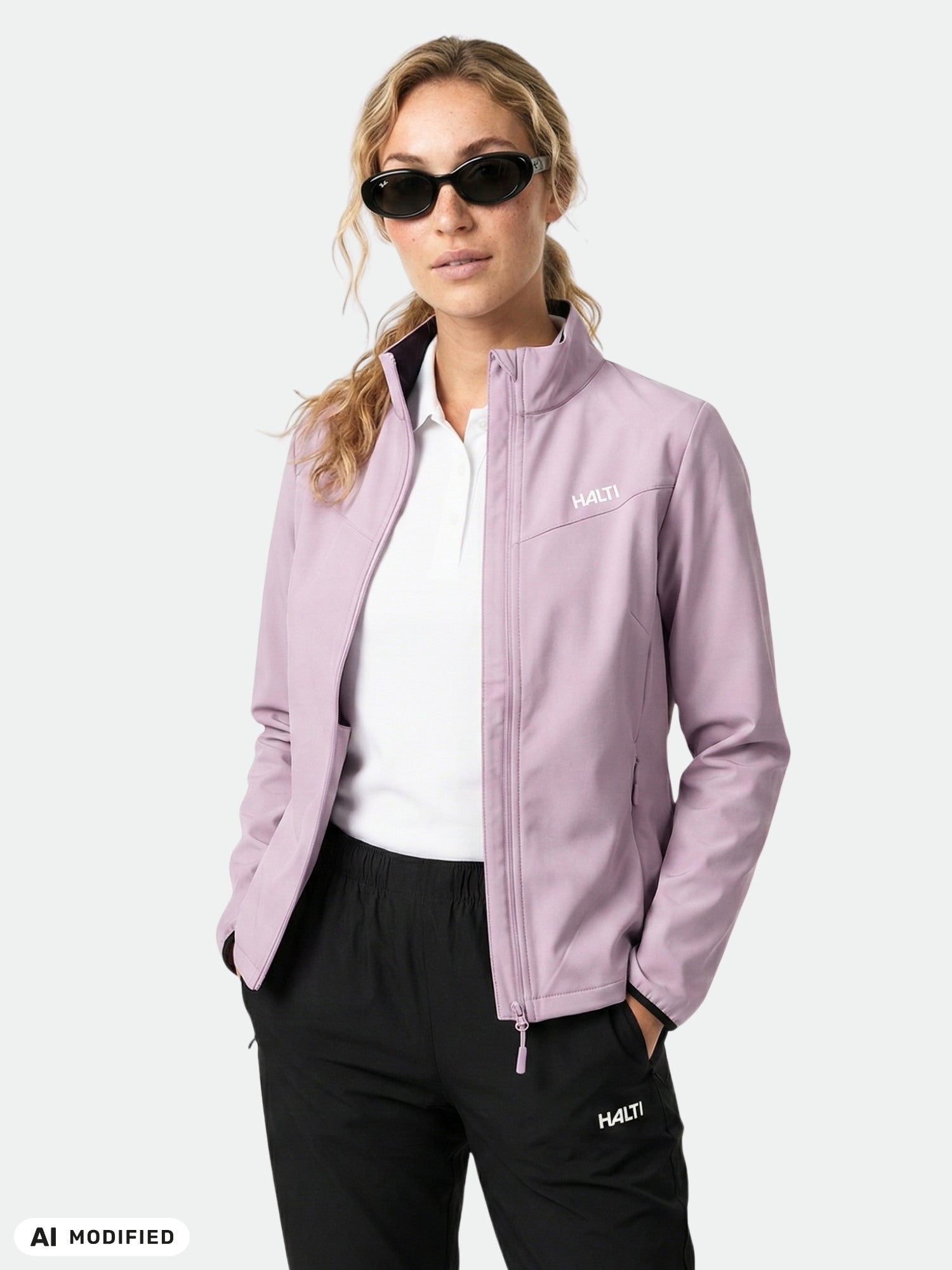 Aurinkolasit päässä oleva nainen on pukeutunut Halti Wedge Softshell -takkiin, valkoiseen paitaan ja mustiin housuihin; hän seisoo kädet taskussa.