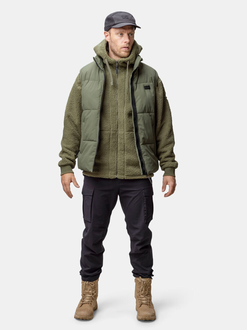 Mies katsoo eteenpäin Halti Penger Classic Puffer Liivi Miesten, vihreä fleece, mustat housut, ruskeat saappaat ja musta lippis.
