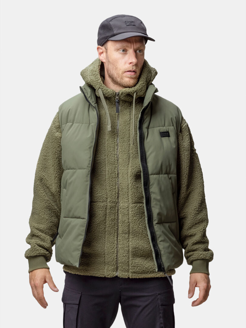 Miehellä on yllään Halti Penger Classic Puffer Liivi Miesten fleecehupparin, mustien housujen ja lippiksen päällä tavallista taustaa vasten.