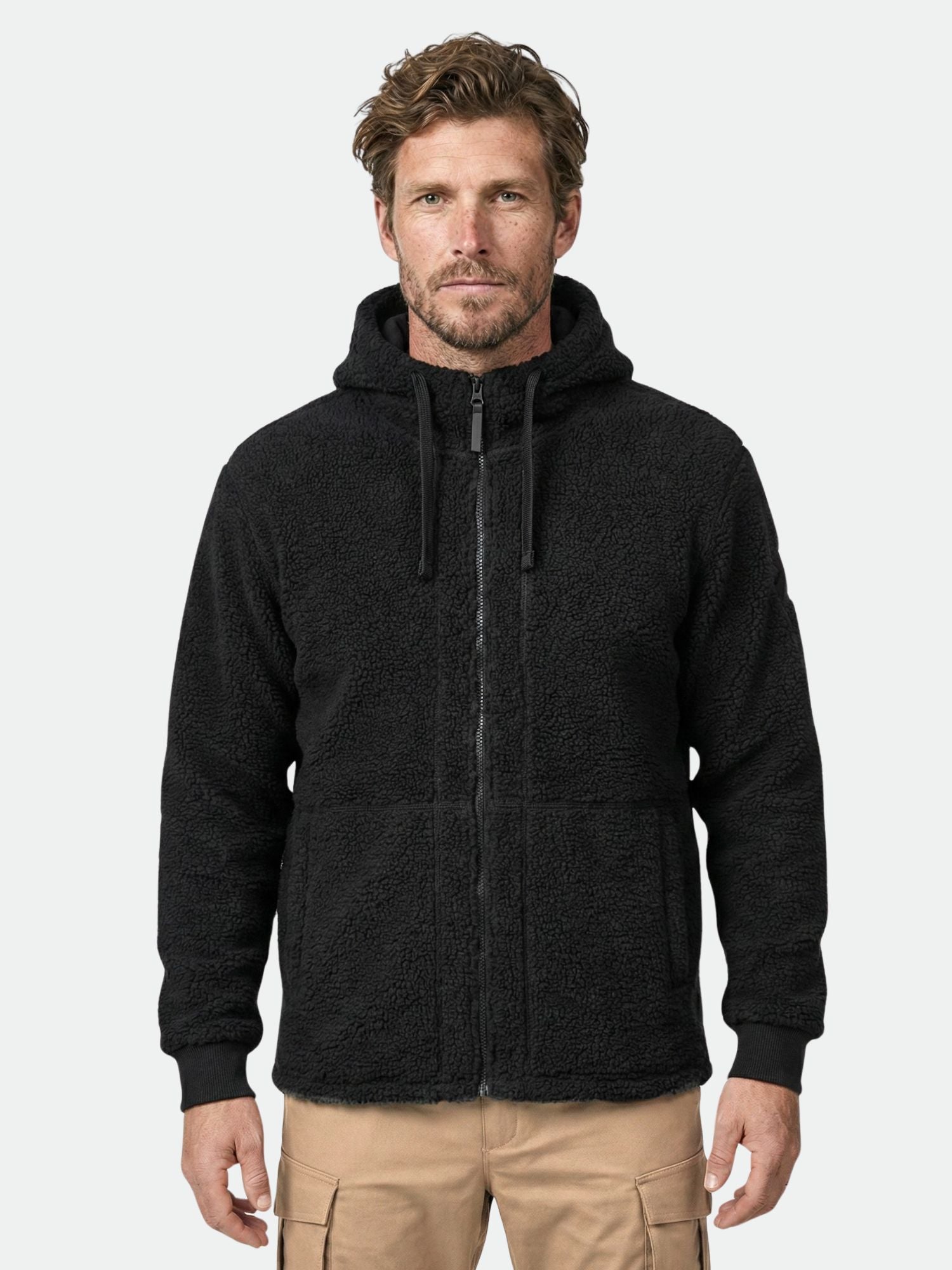 Mies, jolla on Halti Lykky Fleecetakki Miesten ja ruskehtavat rahtitaskuhousut, seisoo vaaleanharmaata taustaa vasten.