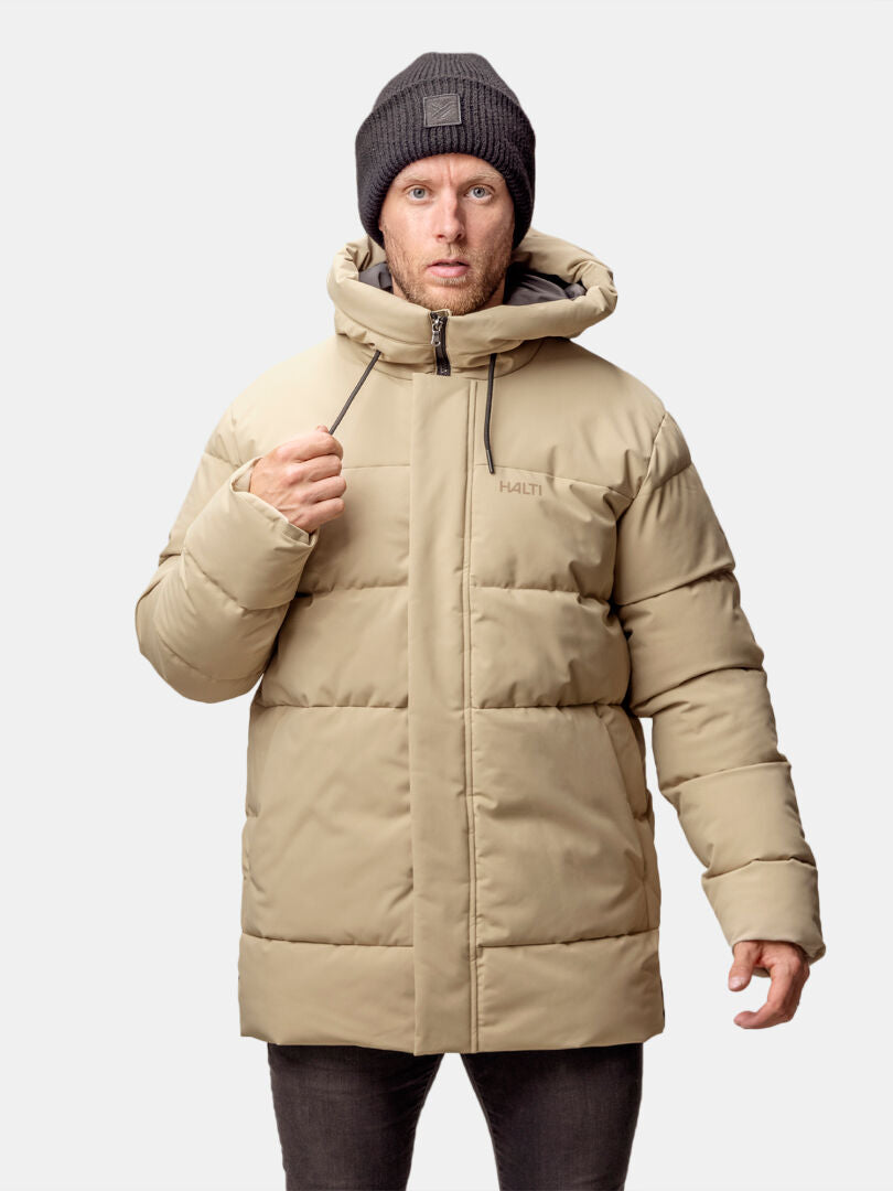 Mies, jolla on beige Halti Sammal Topattu Parkatakki miesten huppu ja tumma pipo, seisoo vaaleaa taustaa vasten.