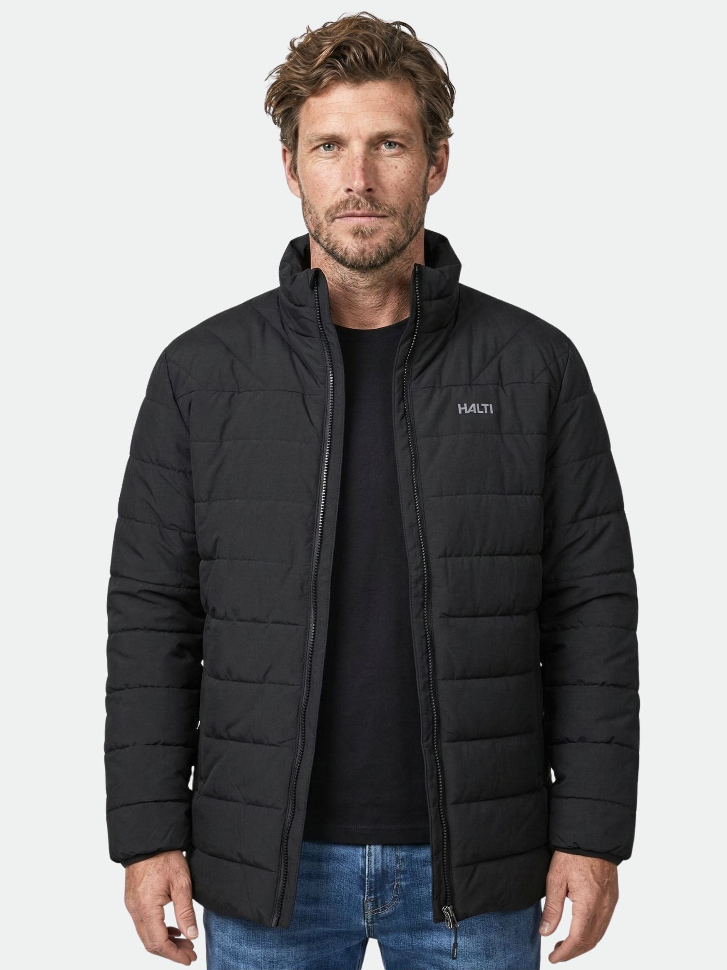 Mustaan Halti Haapa Kevyttoppatakki -miehiin, mustaan paitaan ja sinisiin farkkuihin pukeutunut mies seisoo tavallista taustaa vasten.