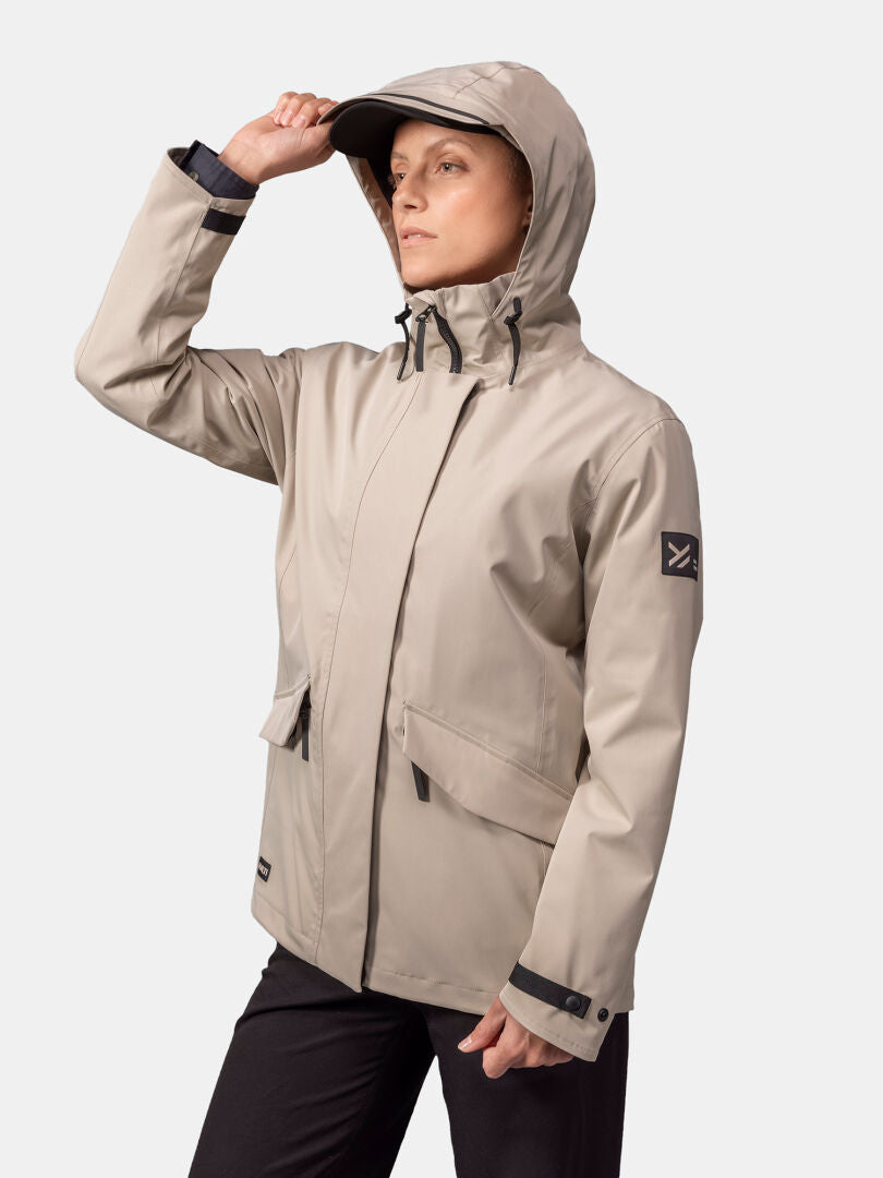 Jollakulla on yllään beige Halti Tokoi DX Takki Naisten ja mustat housut, ja hän seisoo valkoista taustaa vasten.