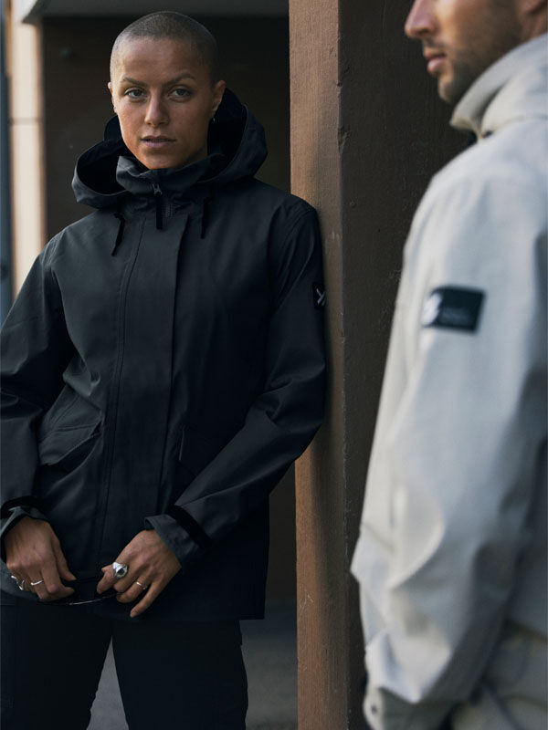 Joku Halti Tokoi DX Takki Naisten -asuun pukeutunut henkilö seisoo seinän vieressä, kun toinen vaaleaan takkiin pukeutunut henkilö kohtaa hänet.