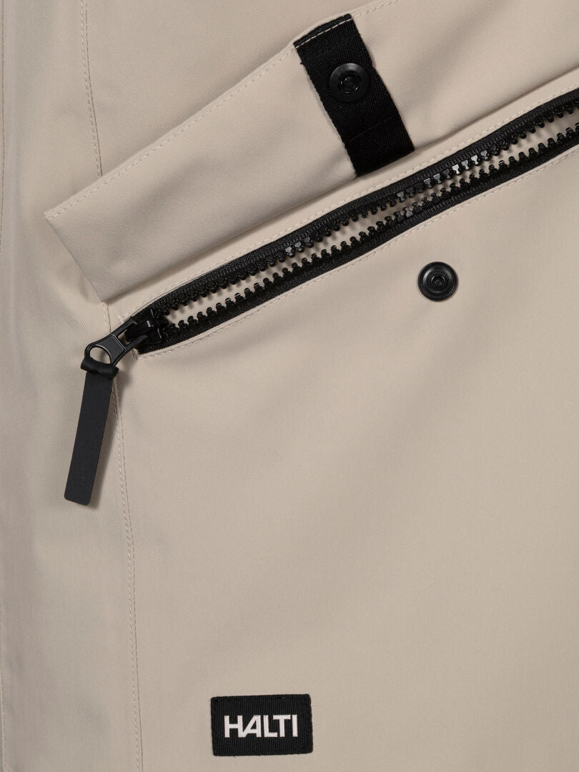 Lähikuvassa Halti Tokoi DX Takki Miesten beige, jossa on vetoketjullinen tasku, napitettu läppä ja musta tuotemerkki helmassa.