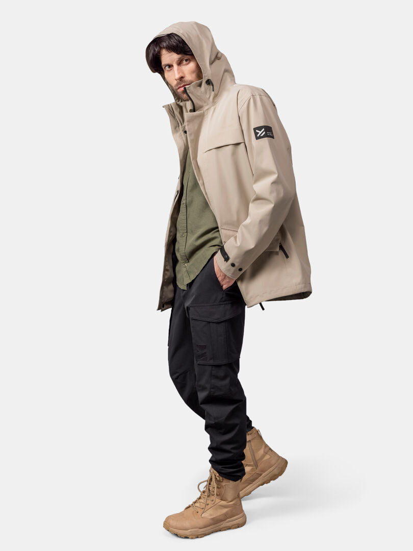 Mies, jolla on beige Halti Tokoi DX takki, vihreä paita, mustat rahtitaskuhousut ja ruskehtavat saappaat tavallista taustaa vasten.