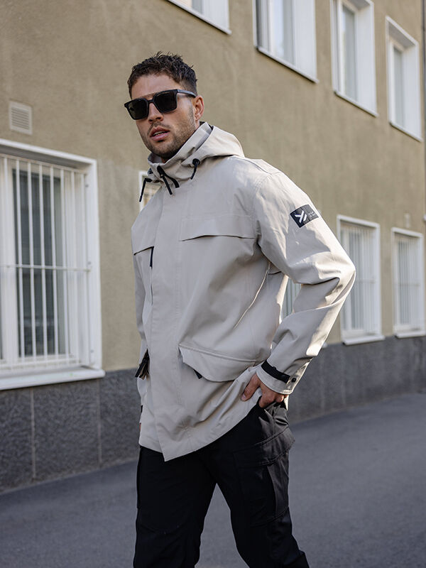 Aurinkolaseihin ja kevyeen Tokoi DX Takki Miesten by Halti -asuun pukeutunut henkilö seisoo jalkakäytävällä lähellä kalteroituja ikkunoita.