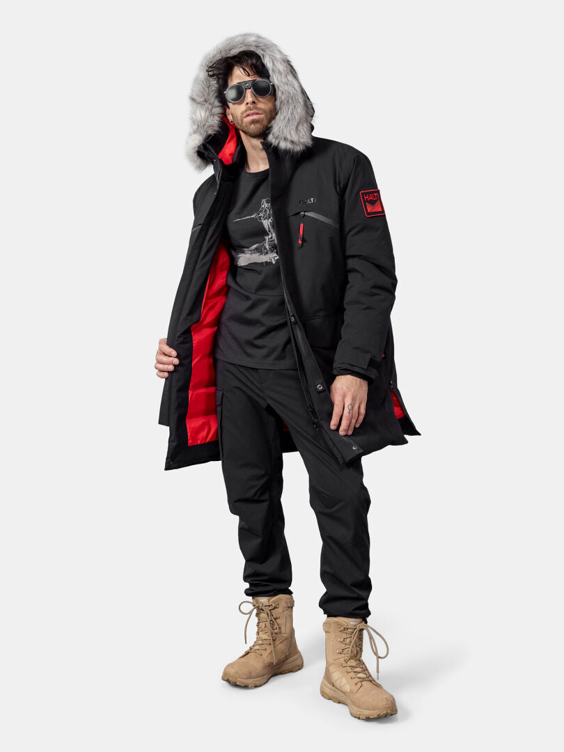 Miehellä on Halti 1976 Down Parka, mustat housut, ruskehtavat saappaat, aurinkolasit ja musta graafinen t-paita.