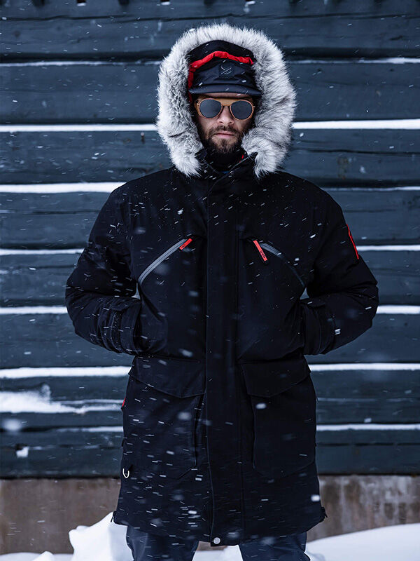 Henkilö, jolla on Halti 1976 Down Parka Unisex, aurinkolasit ja hanskat, seisoo puisen seinän vieressä lumisateessa.