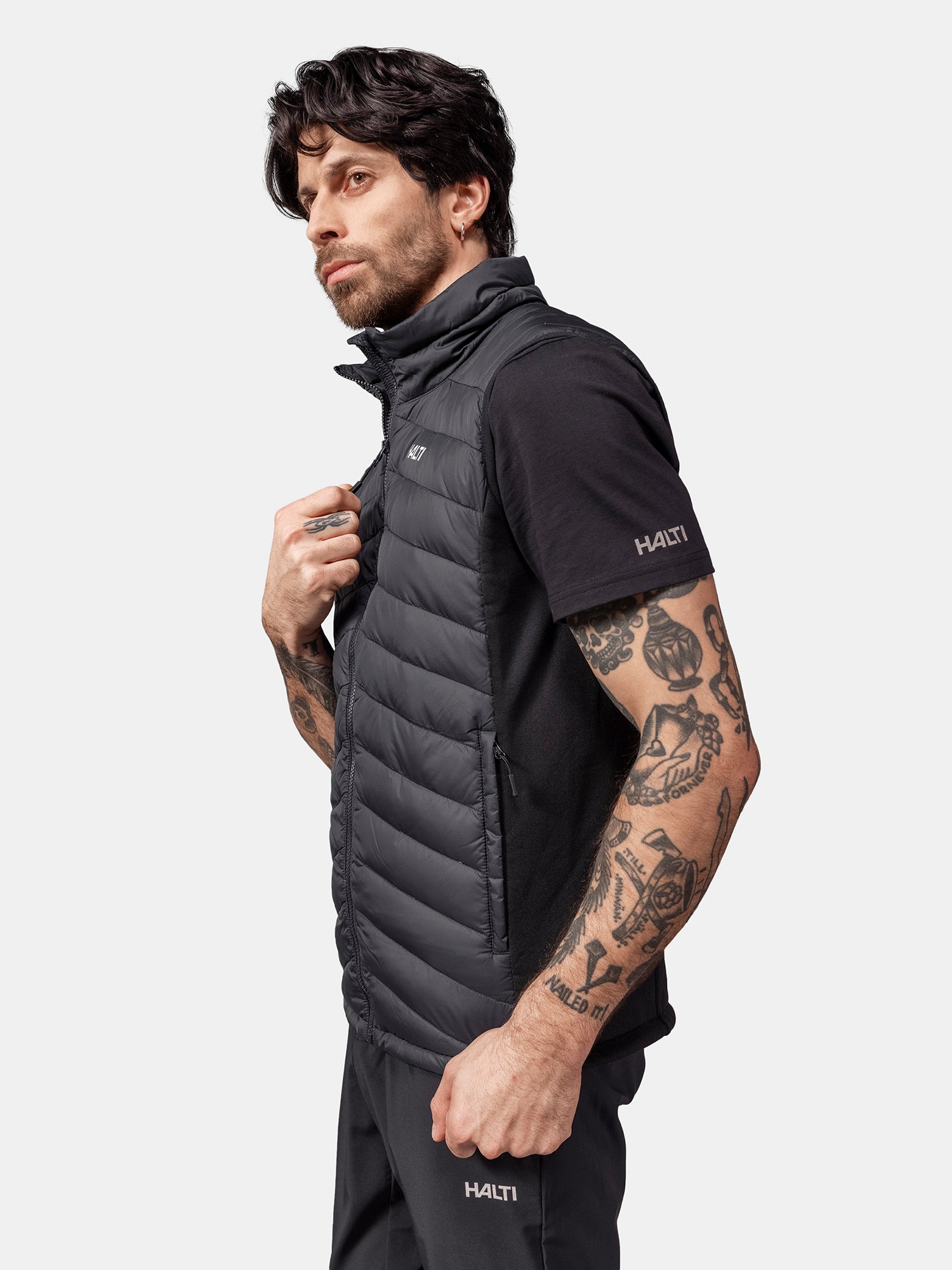Tatuoitu mies pukeutuu Halti Huippu Hybrid Untuvaliiviin, mustaan Pertex Quantum puskuriliiviin, mustan t-paidan ja housujen päälle.