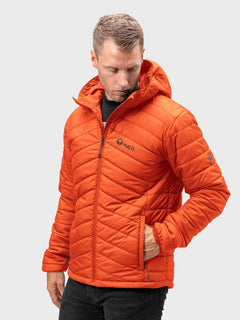 Mies pukeutuu Halti Komu Kevytvanutakki Miesten oranssiin ja mustiin housuihin vaaleanharmaata taustaa vasten.