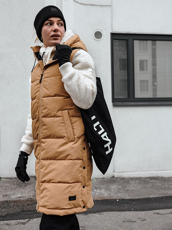 Henkilö pukeutuu ruskehtavaan puskuriliiviin, valkoiseen fleecehuppariin ja Halti Viiri Fleece Hanskatiin kaupungin jalkakäytävällä.