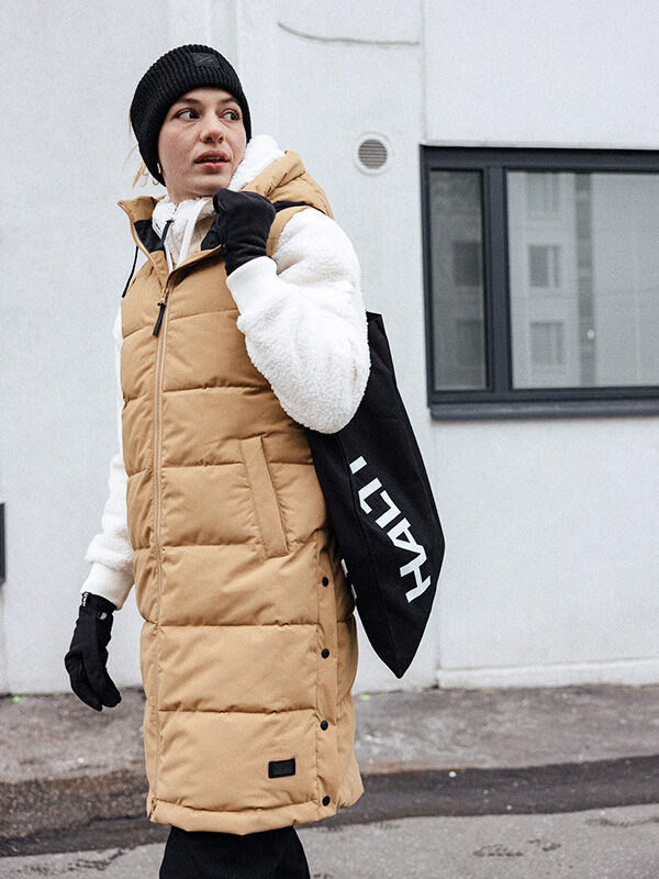 Ulkona oleva henkilö, jolla on ruskeat puskuriliivit, Halti Huurre Panta, valkoinen fleece, käsineet ja musta laukku.