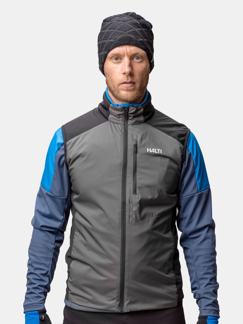 Mies, jolla on harmaa tikattu liivi, sininen paita ja Halti Crostie Merinopipo, valmiina talviliikuntaan tavallista taustaa vasten.