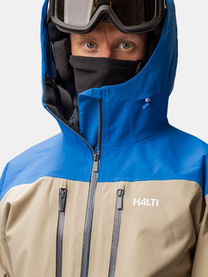 Henkilö, jolla on sininen ja beige Halti-takki, musta naamari, Trone Fleece Tuubihuivi ja hiihtolasit, katsoo kameraan.