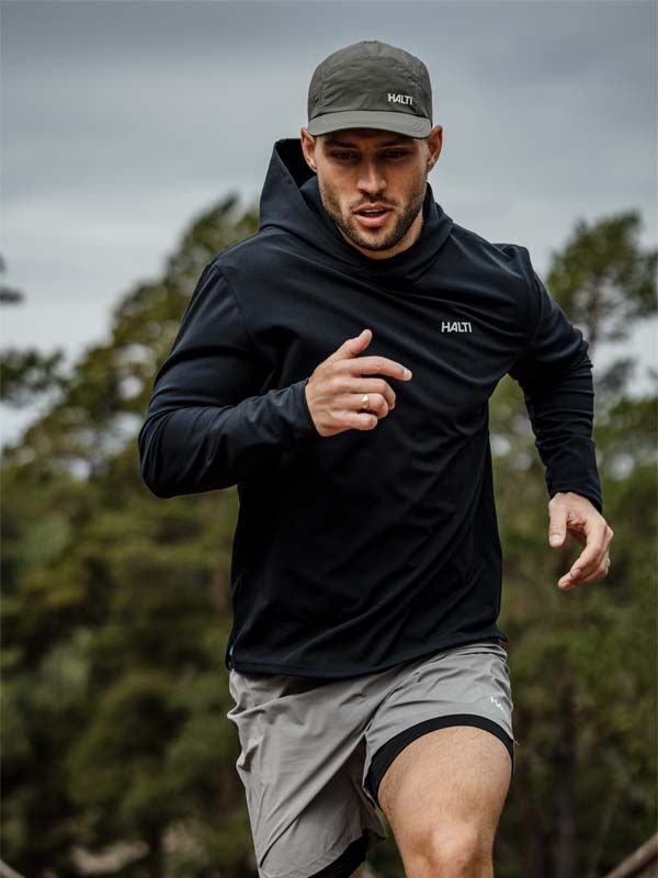 Mies lenkkeilee ulkona mustassa hupparissa, harmaissa shortseissa ja Halti Luovi Lippiksessä, taustalla puita.