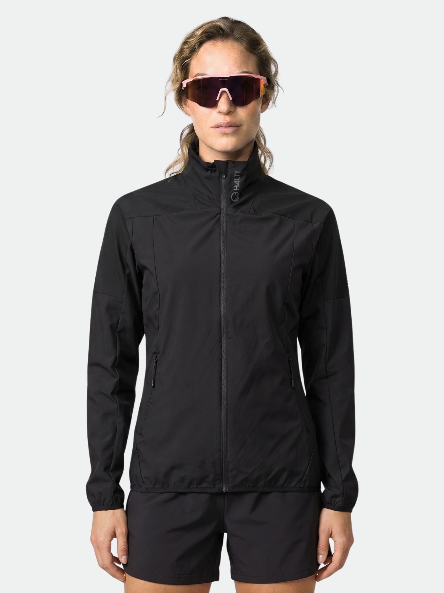 Henkilö, jolla on musta Halti Urbanite Naisten Treenitakki, mustat shortsit ja aurinkolasit, seisoo kasvot menosuuntaan.