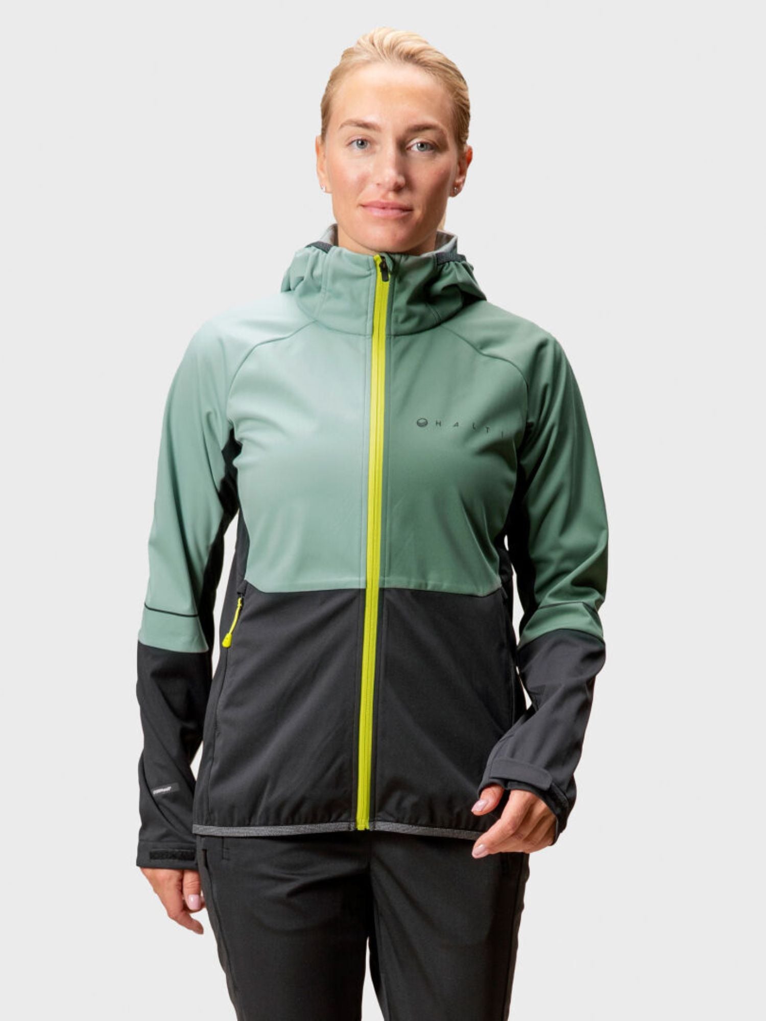 Henkilöllä on yllään Halti Exhale Stormwall Takki Naisten, vihreä ja musta softshell-takki, jossa on keltainen vetoketju.