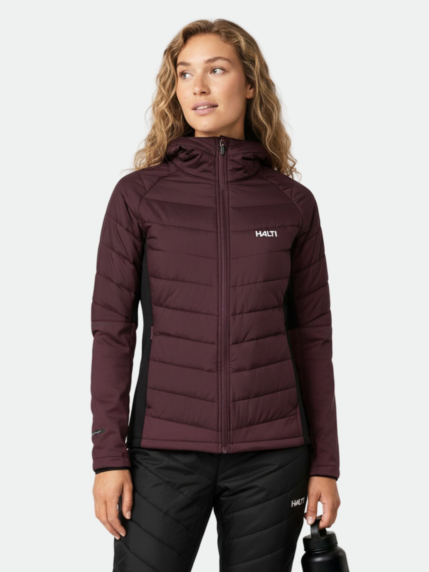 Ruskeankeltaiseen Halti Hanki 2 Hybrid Hiihtotakki Naisten ja mustiin housuihin pukeutunut henkilö pitelee vesipulloa ja katsoo sivulle.