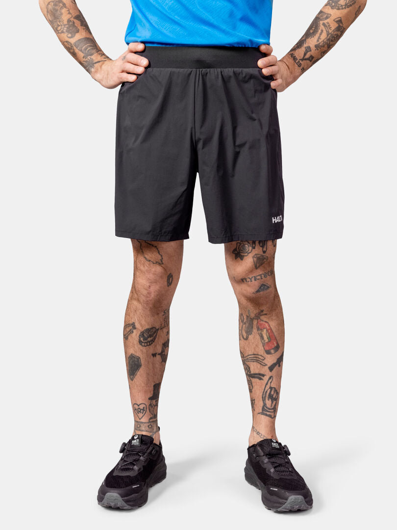 Henkilö seisoo kädet lanteilla Halti Uurto Shortsit Miesten, mustat lenkkarit, sininen paita, näyttää tatuoidut jalat.