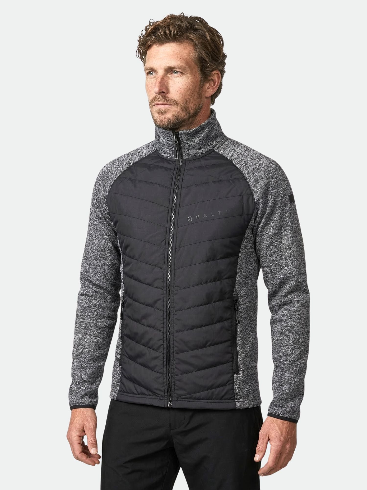 Mies, jolla on Halti Streams Hybrid Välitakki Miesten, jossa on tikattu etuosa ja kuvioidut hihat, seisoo tavallista taustaa vasten.