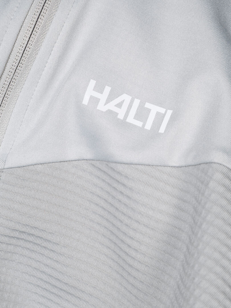 Lähikuva Halti HeatGrid Välitakki Miesten vaaleanharmaasta, vetoketjulla ja valkoisella HALTI-printillä rinnassa.