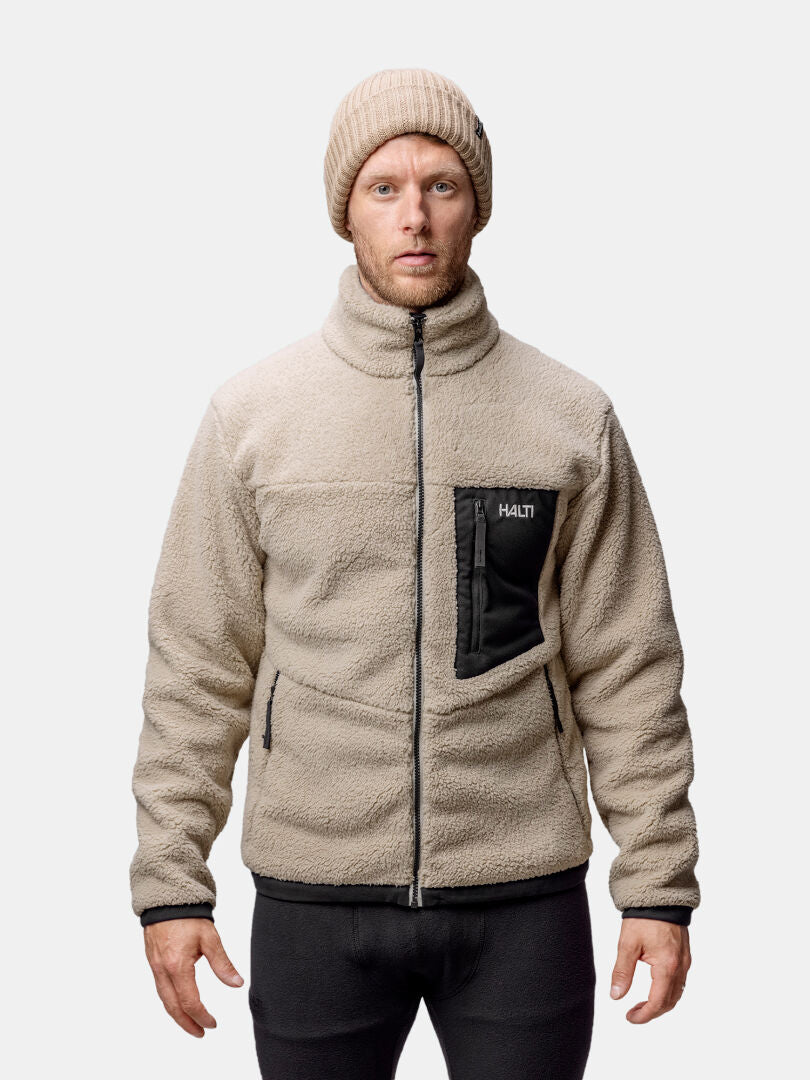Mies, jolla on beige Halti Lumista Polar Fleecetakki Miesten ja neulottu pipo, seisoo vaaleanharmaata taustaa vasten.