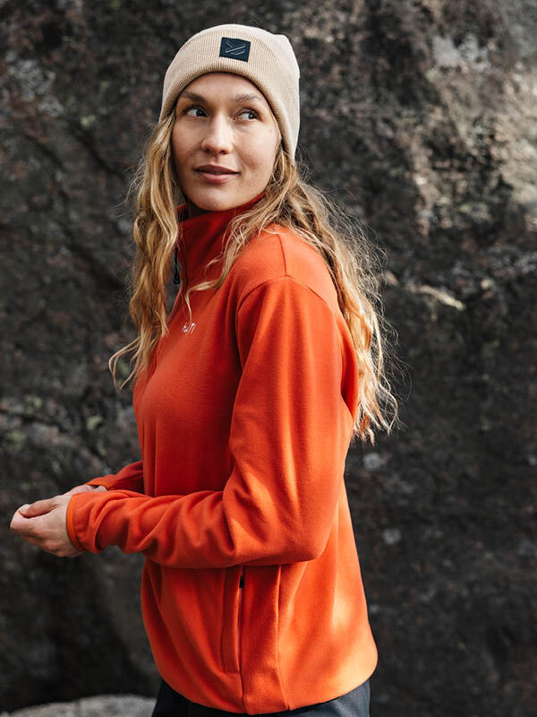 Halti Rockmoon Fleecetakki Naisten oranssiin takkiin ja beigeen pipoon pukeutunut nainen seisoo ulkona ison kiven äärellä.