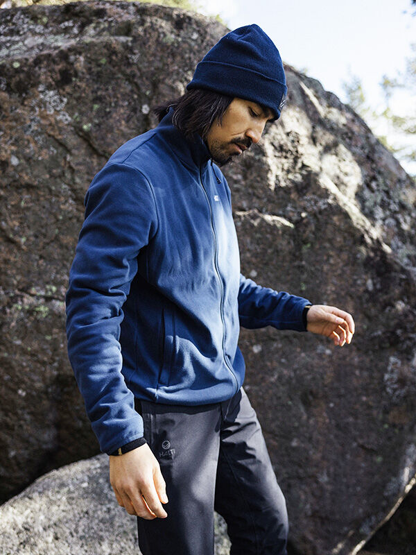 Halti Rockmoon Fleecetakki Miesten ja hattuinen henkilö seisoo ulkona suuren kiven luona katsellen alaspäin kädet ojennettuina.