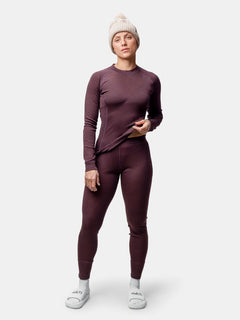 Nainen, jolla on Halti Hossa II Merino Kerrasto -setti, beige neulottu pipo ja valkoiset tossut, seisoo tavallista taustaa vasten.