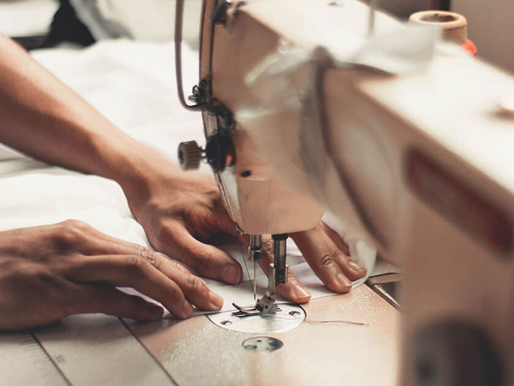 Kaksi kättä ohjaa valkoista kangasta ompelukoneen läpi ja keskittyy ompelun yksityiskohtiin ja koneen osiin.