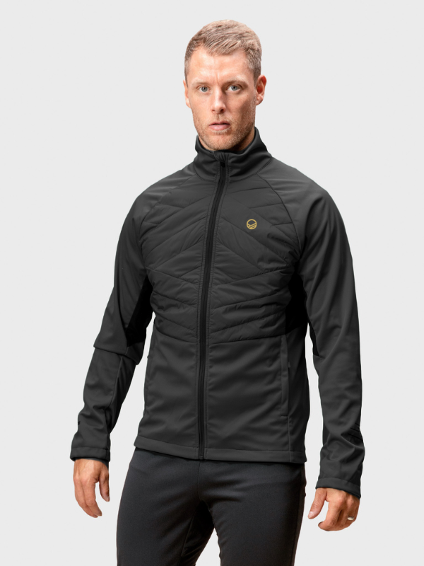 Mies Halti Vinha Hybrid Hiihtotakki Miesten ja mustat housut seisoo tavallista vaaleanharmaata taustaa vasten.